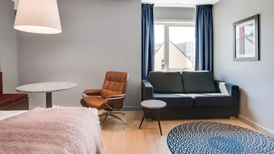 En brun læderstol står i en moderne, minimalistisk stue med en grå sofa, rundt tæppe og marmorbord under en stor pendellampe. Et vindue med blå gardiner i nærheden.
