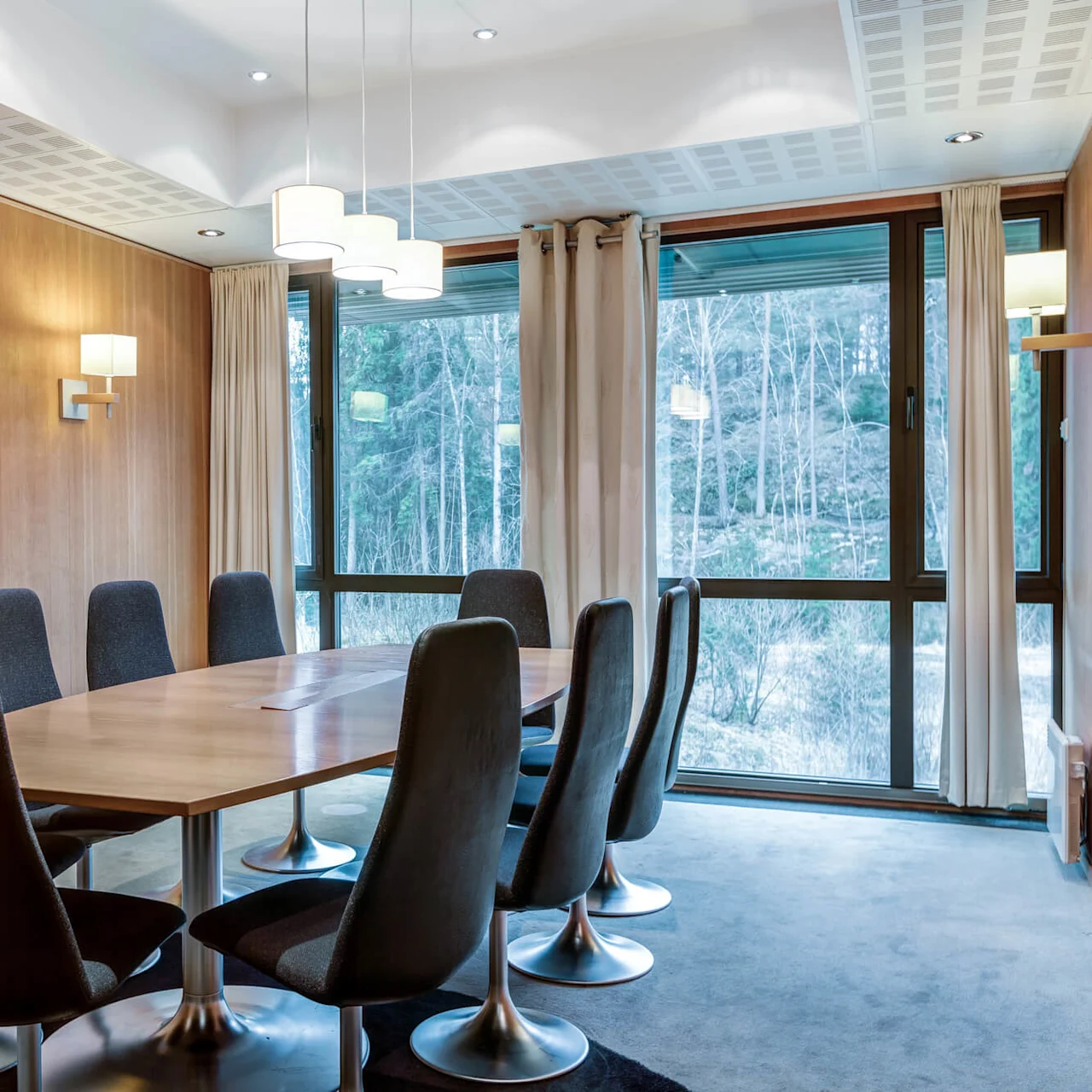 Conference table with ten chairs, surrounded by wood-paneled walls and large windows showing a forest view, under modern pendant lights.