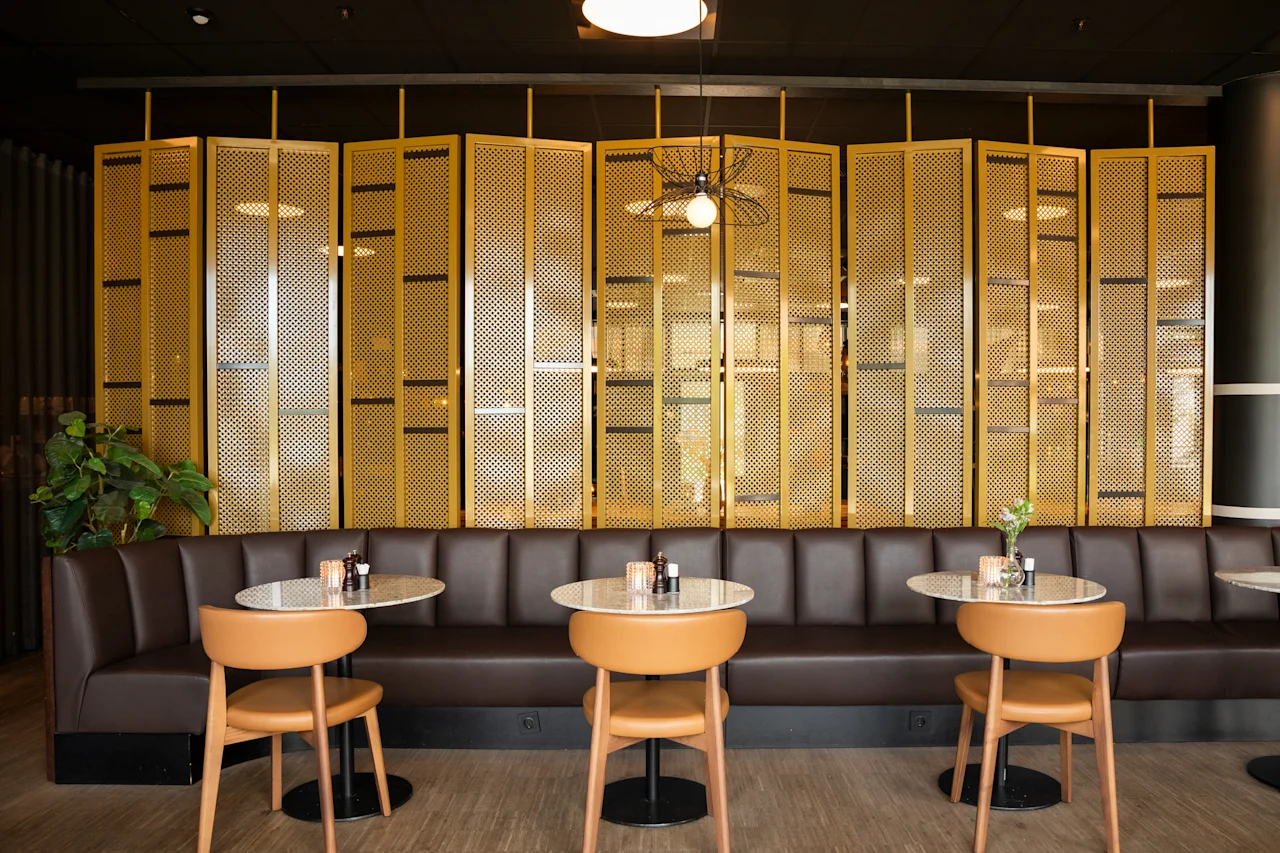 Et koselig restaurantinteriør med en lang brun skinnsofa, små runde bord og en dekorativ gyllen skjerm.