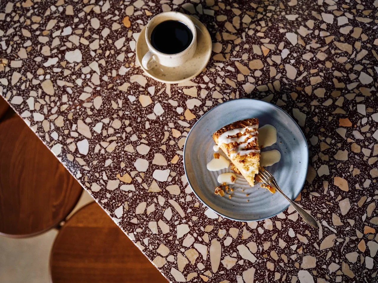 Et stykke kage med glasur ligger på en blå tallerken med en gaffel, placeret på et mønstret stengulv ved siden af en kop sort kaffe.