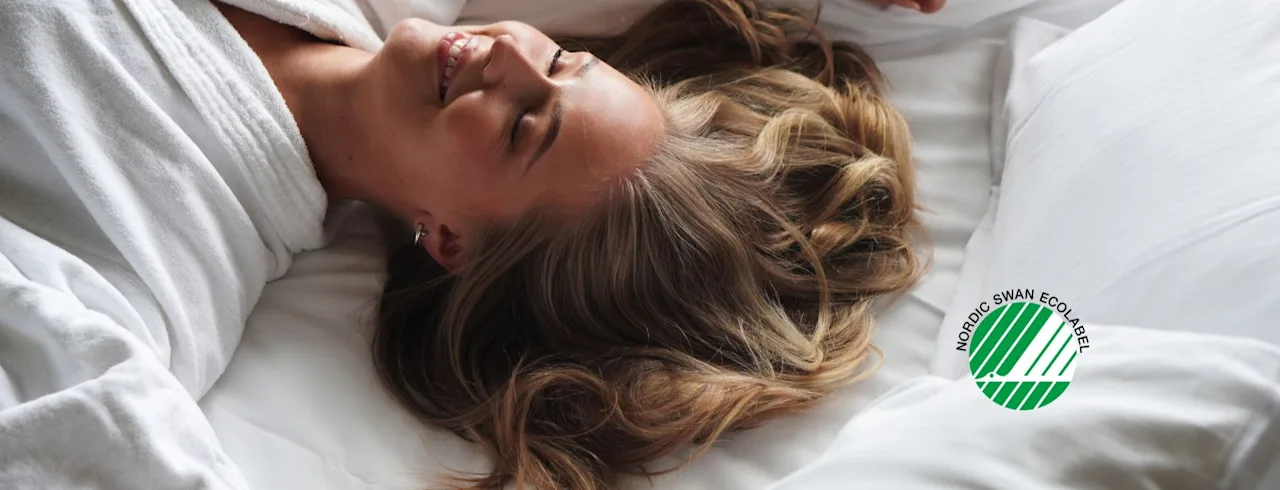 En person kopplar av glatt i en vit morgonrock på en säng med en NORDIC SWAN ECOLABEL kudde, njuter av en lugn stund.