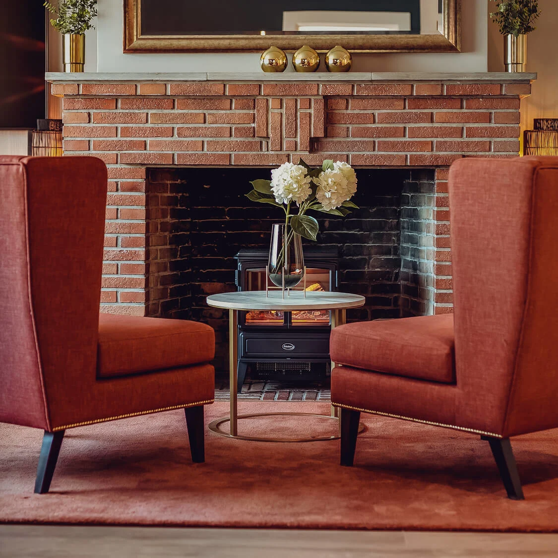 Two terracotta armchairs flanking a round table by a brick fireplace with decorations, in a cozy living room setting.