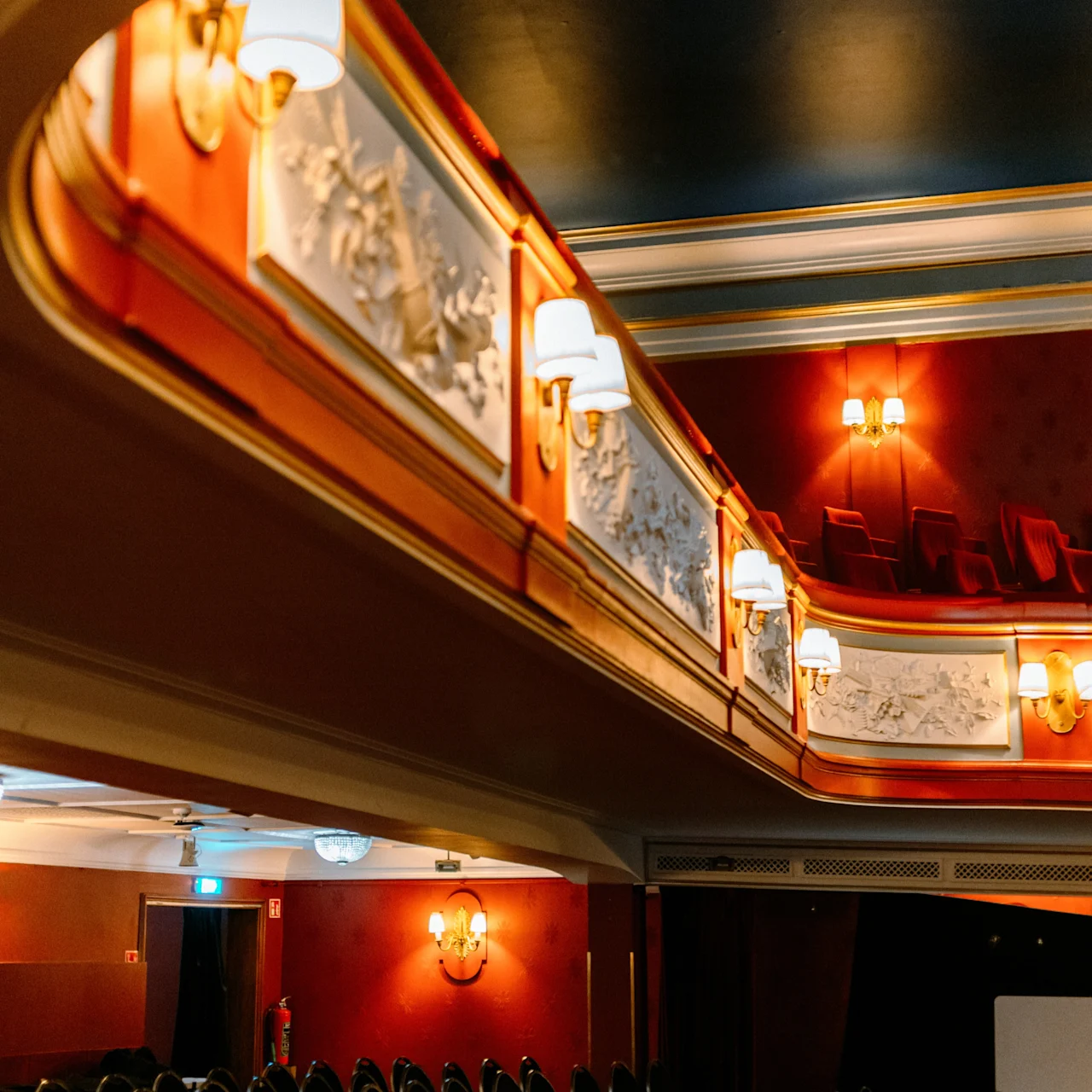 Elegant theater interior featuring rows of empty black chairs. Ornate wall details and glowing wall sconces decorate the red and gold balcony area, creating a warm, inviting atmosphere.