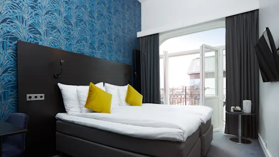 A neatly made bed with white bedding and yellow cushions, against a blue patterned wall, in a room with a balcony door open to the outdoors.