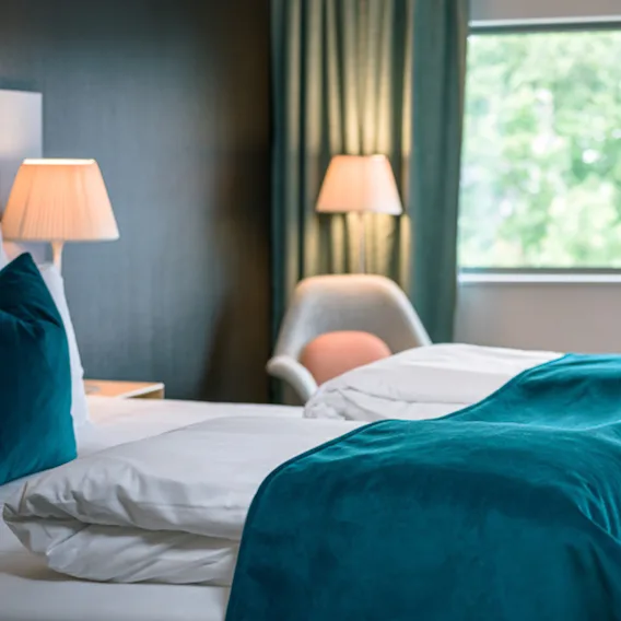 Moderne hotellrom med oppredd seng, mørkegrønt sengeteppe, nattbordslamper og et vindu.