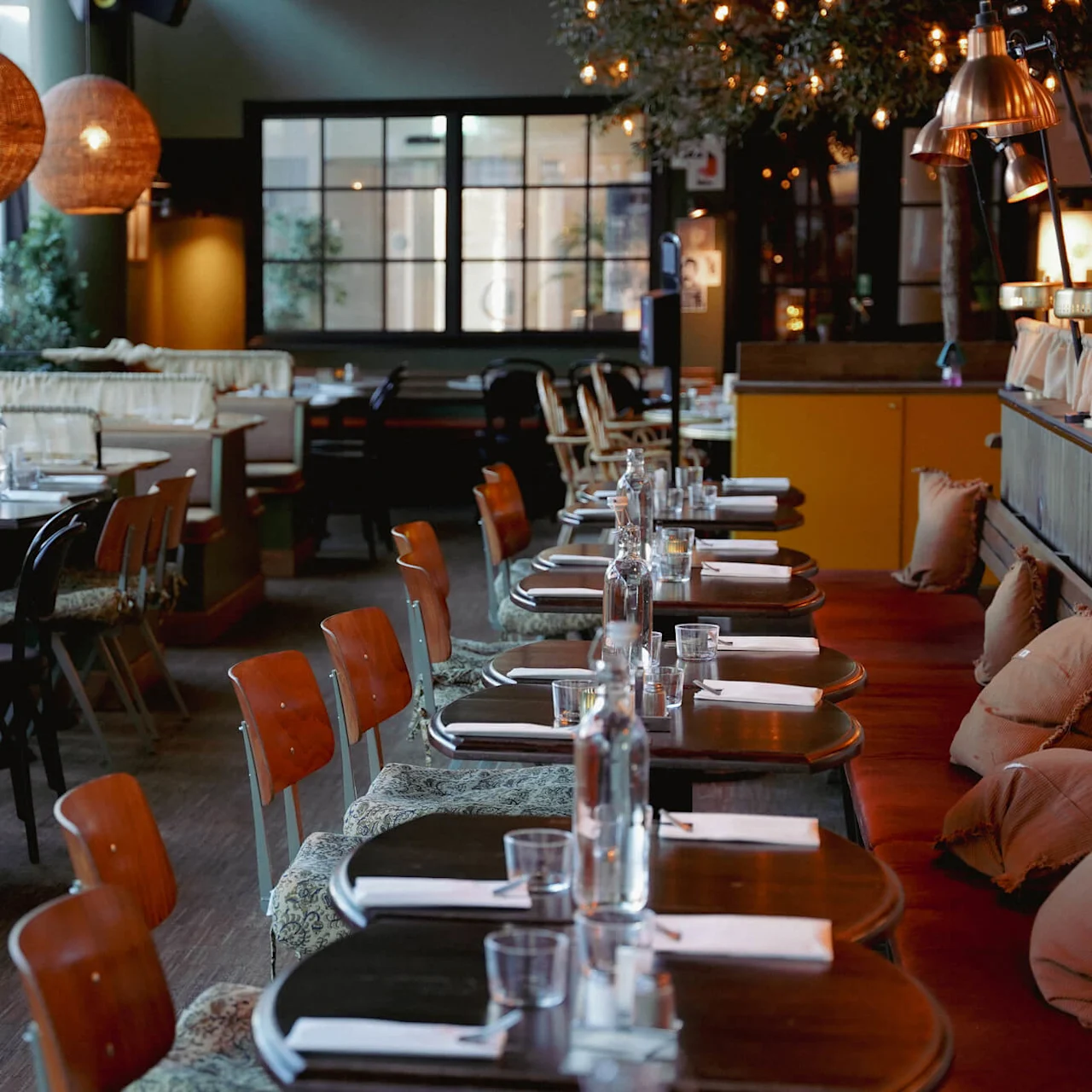Rækker af borde og stole i en dæmpet belyst restaurant, dækket med tallerkener og glas, og pendellamper ovenfor.