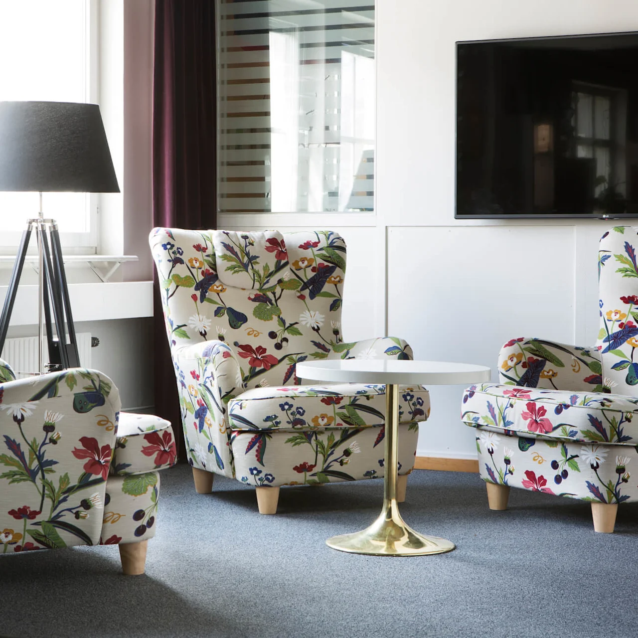 Two armchairs with floral patterns are positioned in a living room, flanking a small round table, with a lamp and a mounted television nearby.