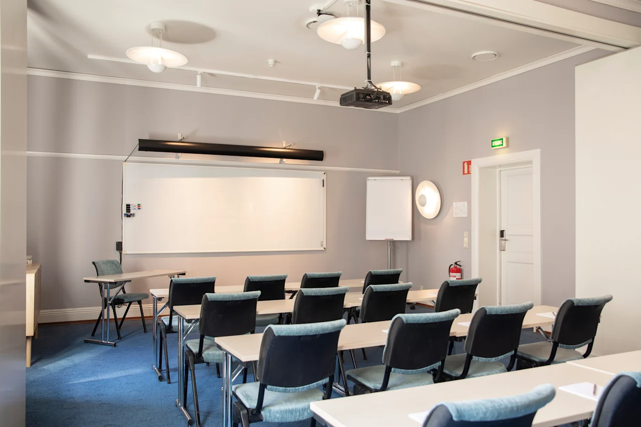Et lyst, tomt konferencelokale med rækker af borde og stole vendt mod en whiteboardtavle og projektor, klar til et møde eller en præsentation.