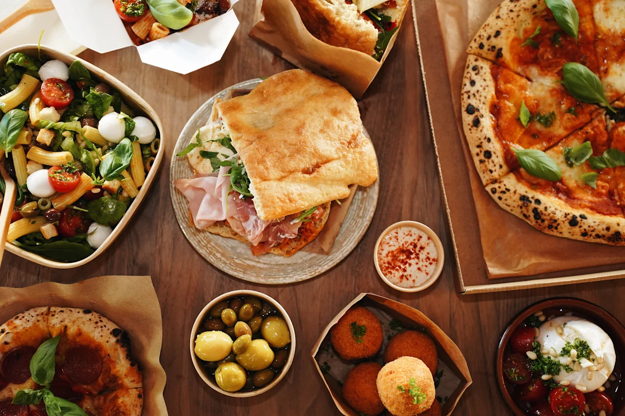 Ett urval av italienska rätter ligger utbrett på ett träbord, inklusive pizza, smörgås, sallad, oliver och arancini, vilket skapar en livfull och aptitretande matsituation typisk för italiensk mat.