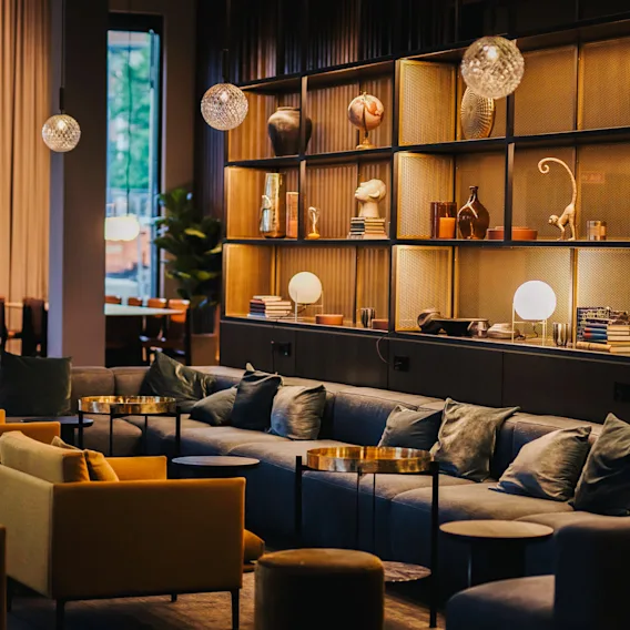 Seating area with blue couches and mustard chairs arranged around small tables; decorative shelves with art pieces and spherical lights illuminate the room's cozy, modern ambiance.