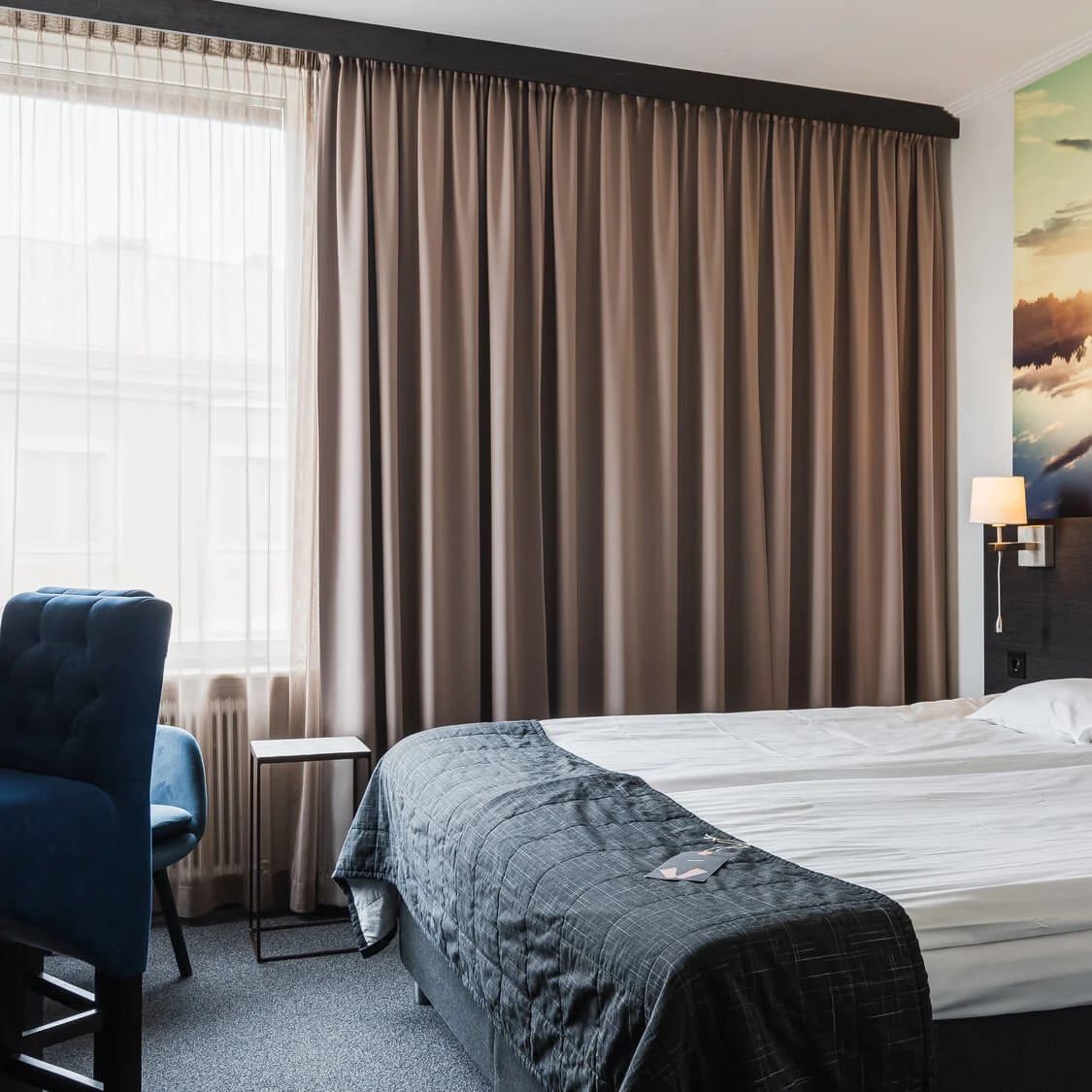 En snyggt bäddad säng med kuddar, i ett hotellrum med en stor väggmålning av en solnedgång över en sjö. En blå stol och ett skrivbord står vid fönstret.