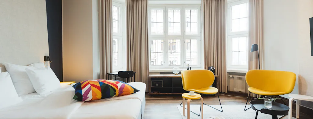 Valoisa ja moderni hotellihuone, jossa on mukava sänky, suuri ikkuna ja viihtyisä oleskelutila keltaisine tuoleineen.