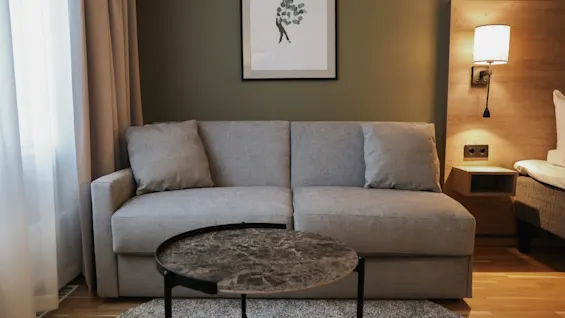A gray sofa sits against a green wall, beneath a framed artwork. A round, marble-topped coffee table is in front, and a lit lamp illuminates the space.