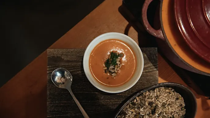 En skål cremet orange suppe med frø og en urtepynt, ved siden af en ske og en skål med frø på et træbord.