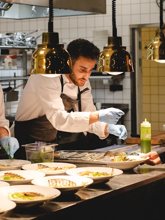 Chefs preparing dishes on a counter, wearing aprons and gloves, in a professional kitchen with hanging lamps and tiled walls. Plates arranged in a row, each with plated food.