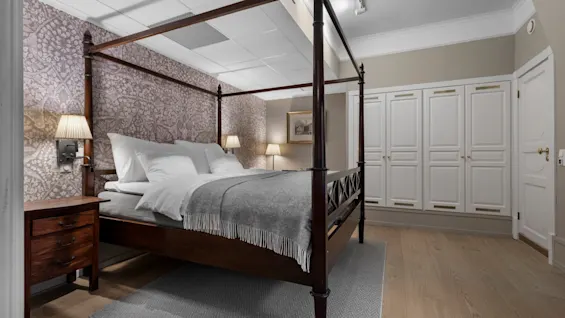 A cozy bedroom featuring a dark wood four-poster bed with white bedding and a grey throw, set against patterned wallpaper.
