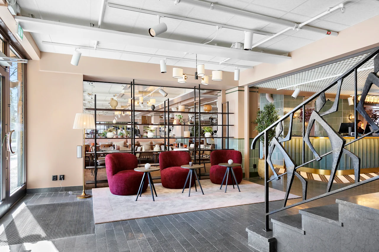 Moderne hotellobby med tre røde lenestoler, små bord og en trapp med dekorativt rekkverk.