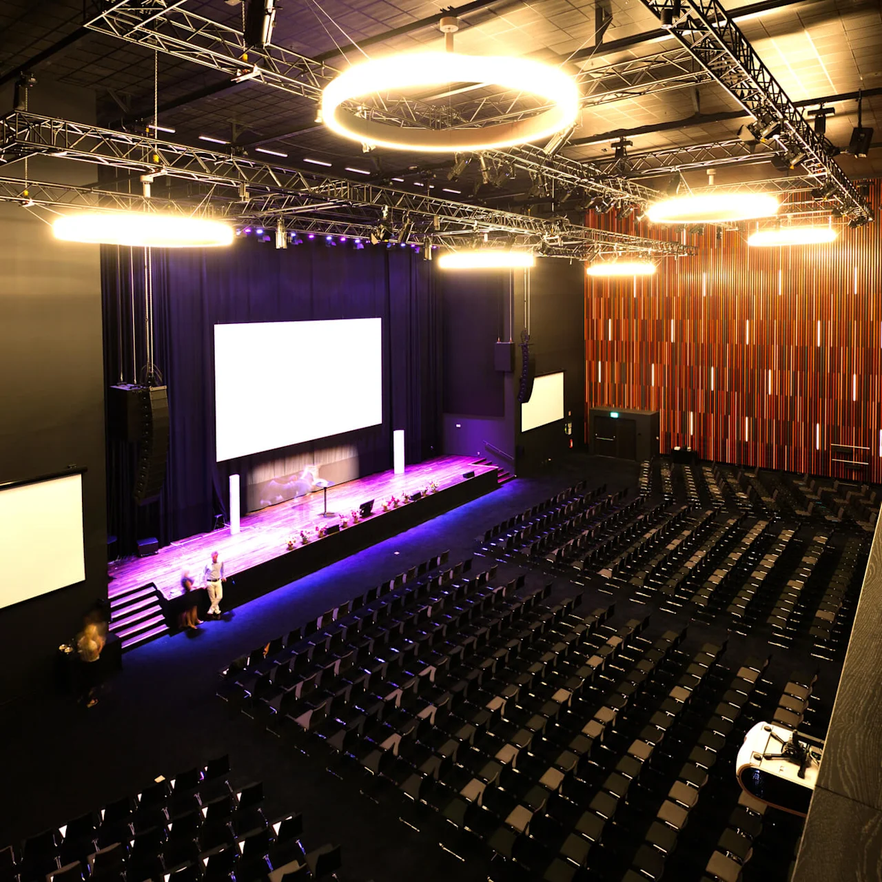 Et auditorium med rader av tomme sorte stoler, scene med store hvite skjermer, og glødende taklys. Veggene er dekorert med vertikale røde og brune paneler.