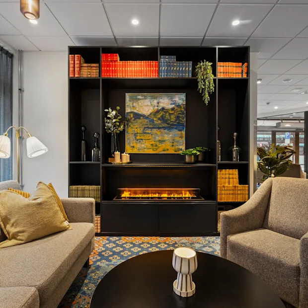 En mysig hotellounge med en bekväm sittgrupp, ett soffbord och en stilren bokhylla med en varm öppen spis.