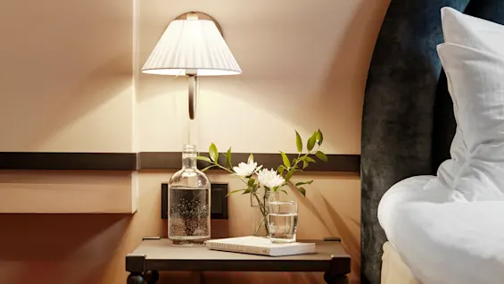 Nightstand holds a lamp, glass water bottle, vase with flowers, beside a bed with a dark headboard in a softly lit bedroom.