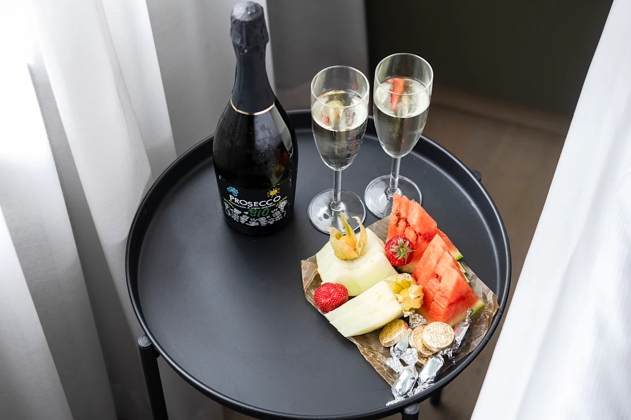 Et lite bord dekket for feiring, med en flaske prosecco og to glass fylt med bobler. Et fargerikt fat med fersk frukt og snacks gir en følelse av luksus.