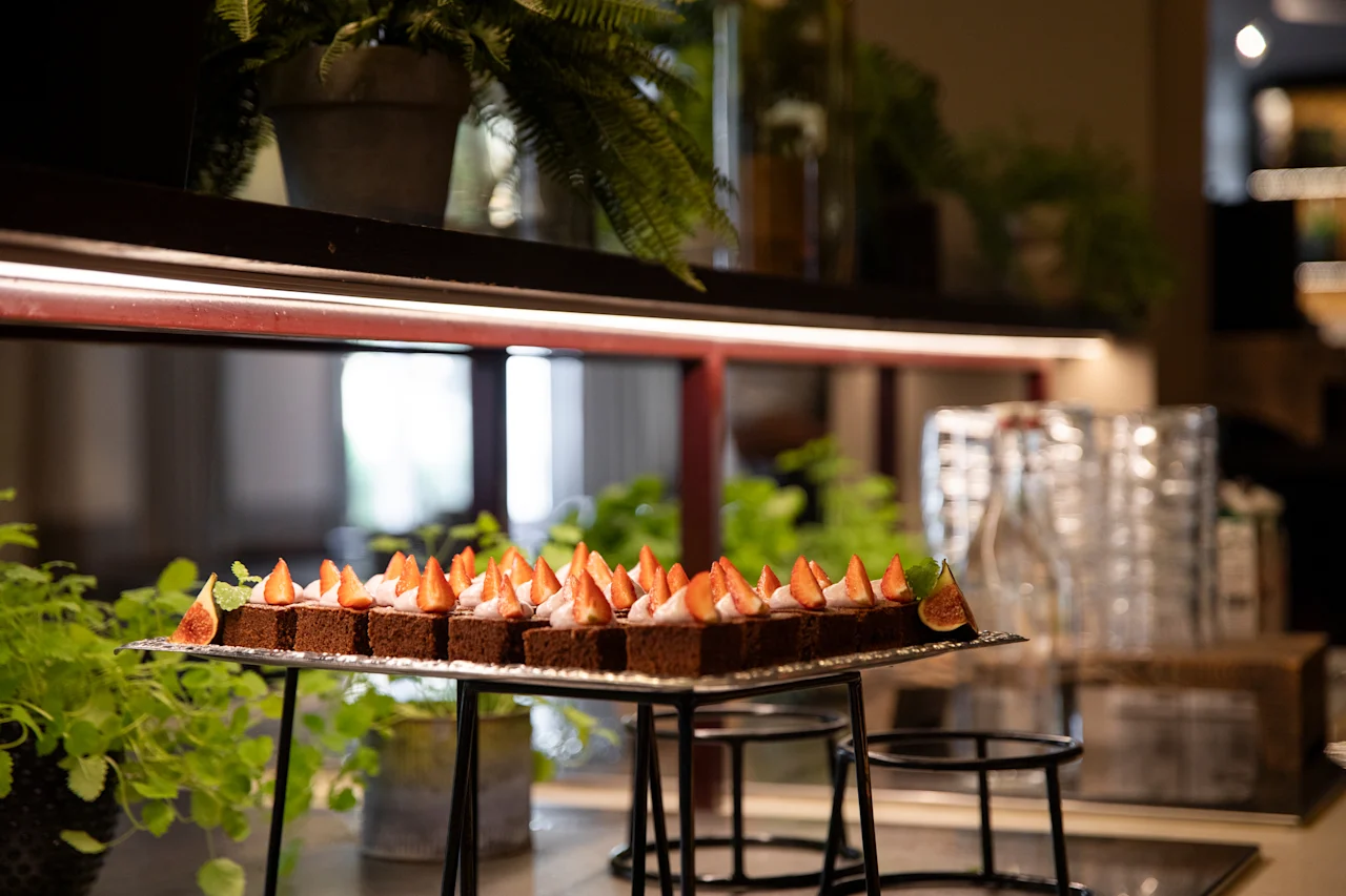 En bricka med chokladkakor toppade med jordgubbar och grädde, presenterade på ett buffébord med gröna växter i bakgrunden.