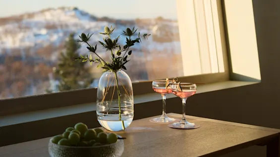 Drikkevarer og druer på et bord ved et vindue, med en klar udsigt over sneklædte bjerge i det fjerne.