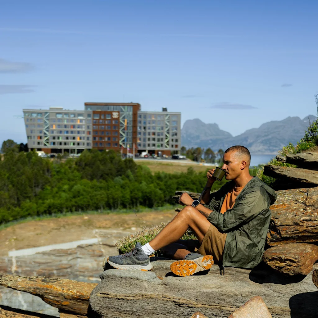 En person sidder på stenet terræn og spiser et måltid udendørs. I baggrunden ses en moderne bygning og bjerge, omgivet af et frodigt landskab under en klar blå himmel.