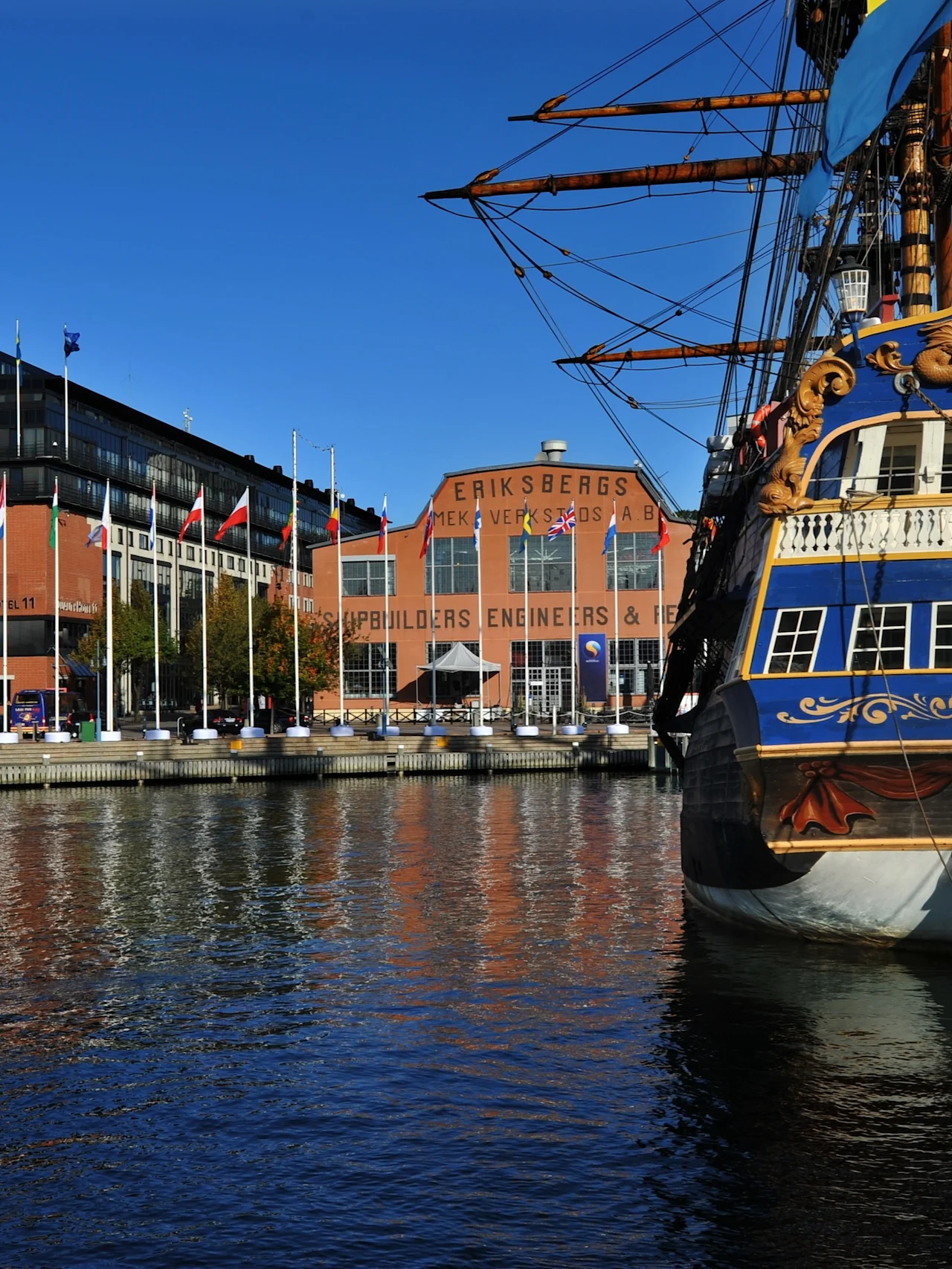 Ett storslaget skepp, GOTHEBORG, med blå och gul akter förtöjt i en hamn. Byggnader i bakgrunden lyder SHIPBUILDERS ENGINEERS.