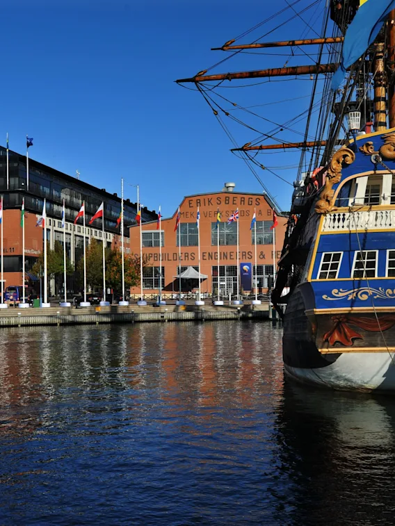 Ett storslaget skepp, GOTHEBORG, med blå och gul akter förtöjt i en hamn. Byggnader i bakgrunden lyder SHIPBUILDERS ENGINEERS.