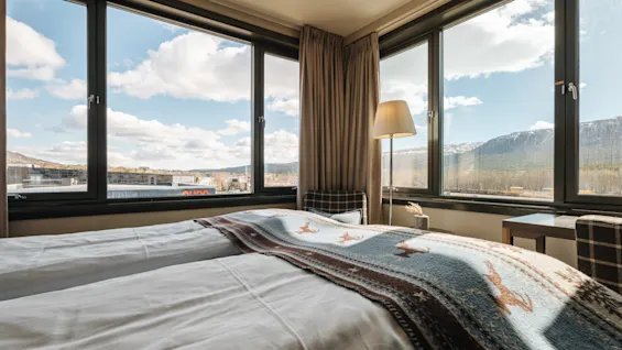 Ljust hotellrum med en stor säng och stora hörnfonster som visar en vacker utsikt över berg och en blå himmel.