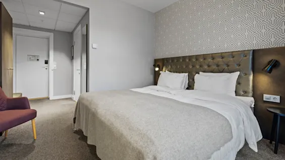 Ett modernt hotellrum med en stor säng, en bekväm fåtölj och stilren mönstrad tapet, vilket skapar en välkomnande atmosfär.