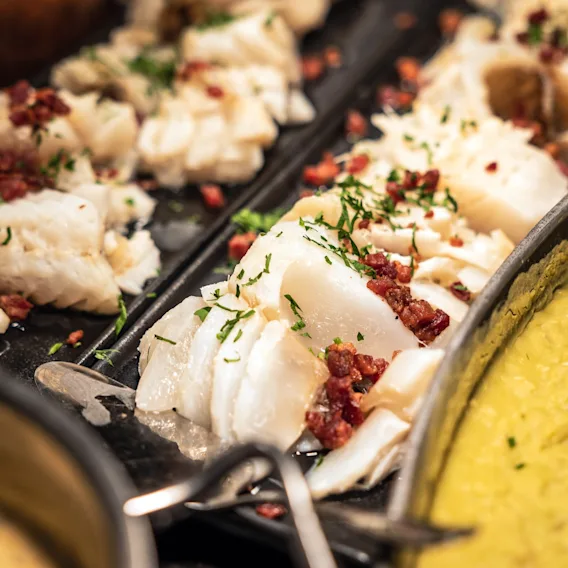 Skiver av kokt fisk toppet med urter og terninger av rødt tilbehør ligger på et fat. I nærheten gir en kremet gul rett fargekontrast til matpresentasjonen.