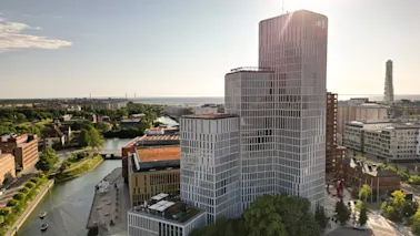 En hög, modern byggnad med rutnätsfasad står vid en kanal i en stadsmiljö med avlägsen utsikt över havet och en bro.