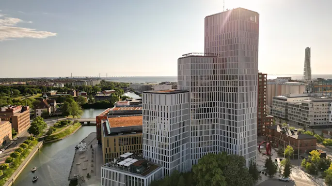 A tall, modern building with a grid-like facade stands by a canal in a city environment with a distant view of the sea and a bridge. 