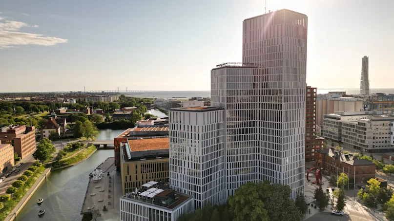 A tall, modern building with a grid-like facade stands by a canal in a city environment with a distant view of the sea and a bridge. 