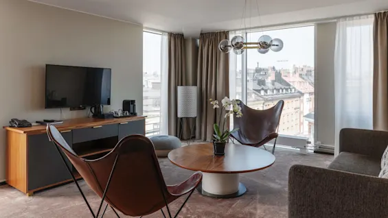 Ett välupplyst, modernt vardagsrum med ett runt bord, stolar, en tv och stora fönster med utsikt över staden.