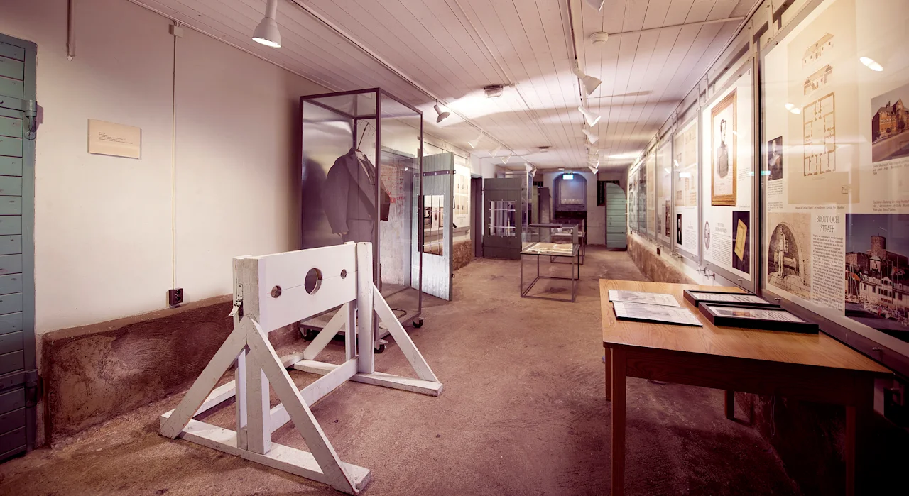 A historical museum exhibit in a long hallway with a wooden stockade, display cases, and framed documents on the wall. Visible text: BRITTISK STRAFF.
