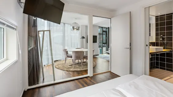 A modern bedroom with an open sliding door leading to a dining area, sunlight streams in, highlighting the clean, contemporary design.
