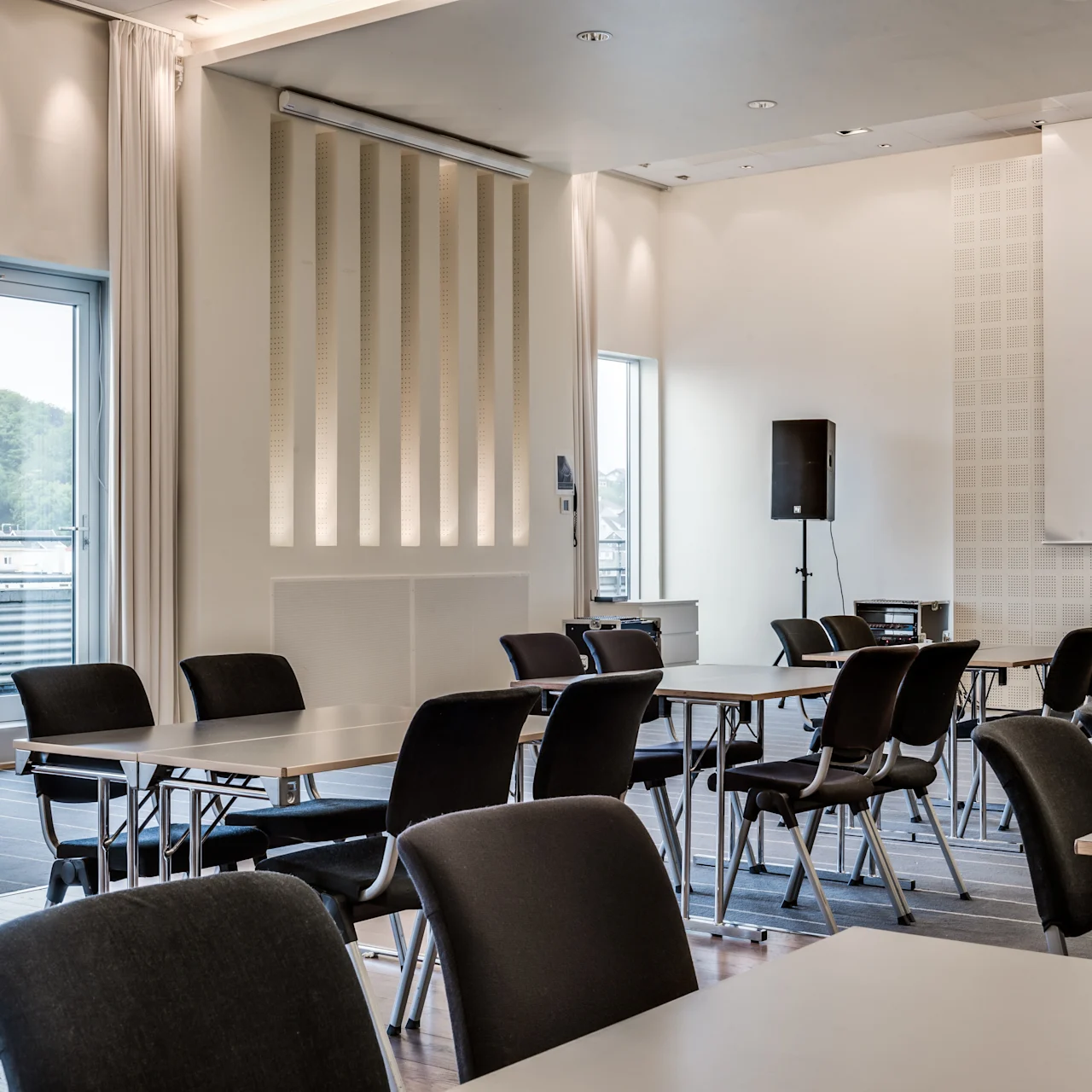 Pöydät ja tuolit on järjestetty konferenssitilaan, jossa on luonnonvaloa ikkunoista. Tekstiä ei ole. Iso näyttö ja äänilaitteet viittaavat esitysjärjestelyihin.