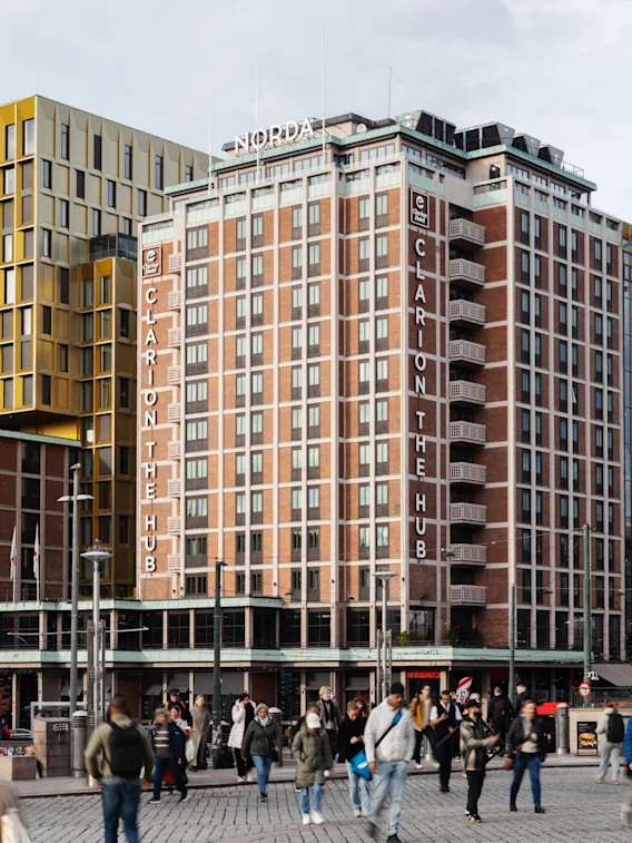 People walk on a city street in front of a tall building with CLARION THE HUB written on its side, surrounded by other urban structures.