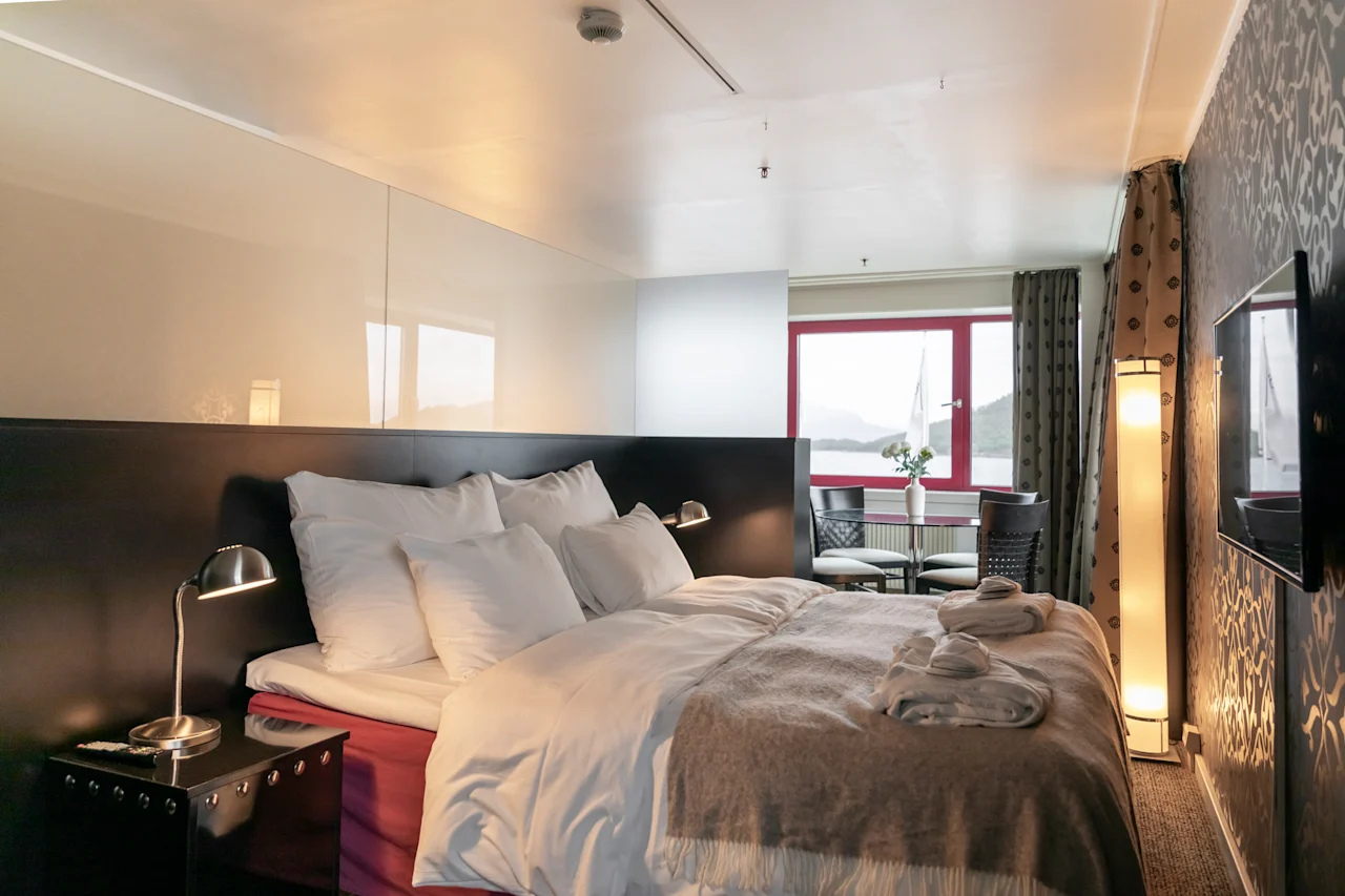 Viihtyisä hotellihuone, jossa on mukava sänky, ruokailutila ikkunan äärellä ja luonnonkaunis näkymä järvelle ja vuorille.