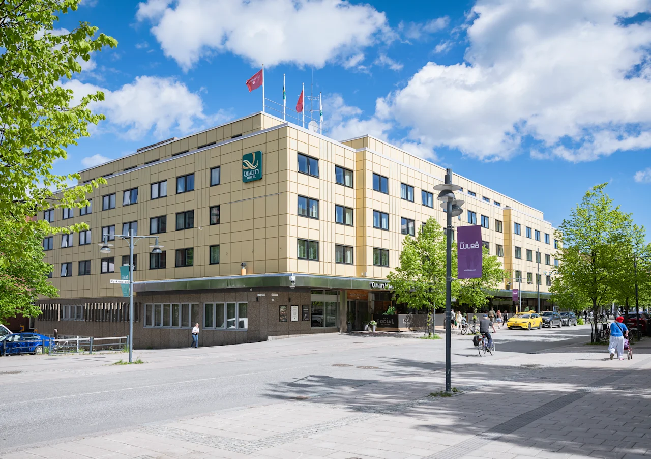 En stor hotellbyggnad med en grön logotyp under blå himmel, med människor som går och cyklar framför.