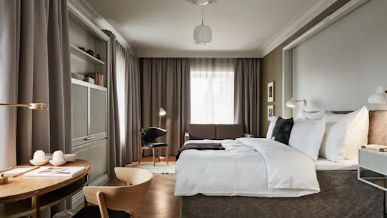 En pent, oppredd seng står på et romslig hotellrom med nøytral innredning, tregulv og møbler. Gardiner dekker vinduene, og et skrivebord med stoler er i nærheten.