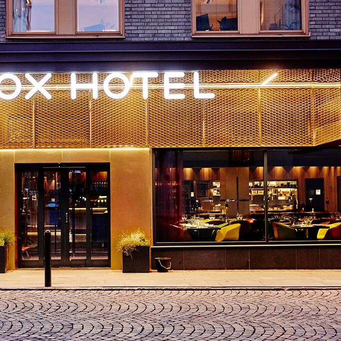 VOX HOTEL-skiltet lyser klart på en bygningens facade. Nedenfor fører glasdøre ind i et varmt oplyst interiør med spiseborde. Brostensbelagt gade foran.