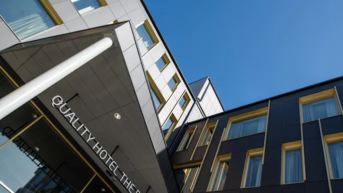 Fasaden på en moderne bygning med store vinduer; inngangen viser "QUALITY HOTEL THE BOX" i klare bokstaver. Himmelen er klar blå og fremhever bygningens geometriske design.