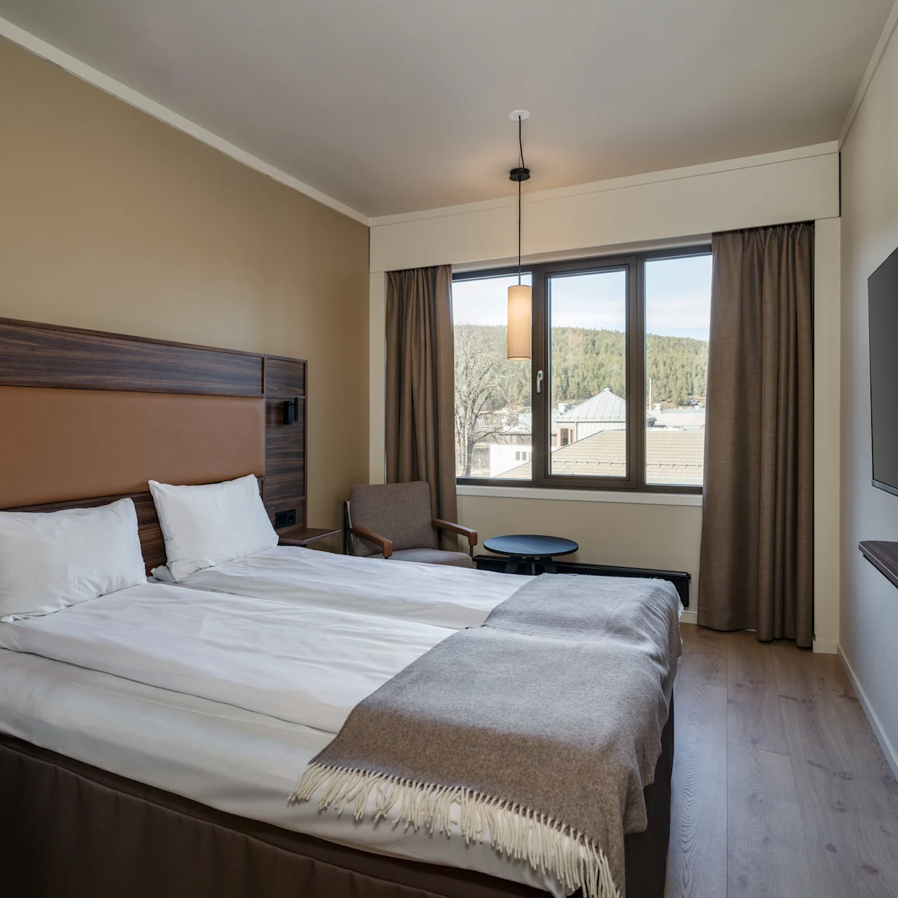 A neatly made bed with a brown headboard, positioned in a hotel room. Natural light enters through a large window with curtains, beside a small seating area and a wall-mounted TV.