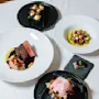 Et udvalg af gourmet retter præsenteret på elegante tallerkener, med kød, grøntsager og en lyserød dessert, arrangeret på en hvid dug.
