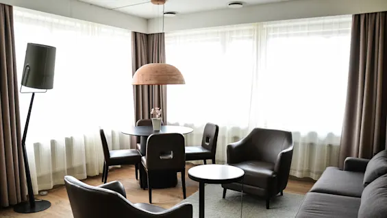 A living room features a small round table with a potted plant, surrounded by chairs. Armchairs and a sofa provide seating. Soft lighting and curtains complete the cozy setting.