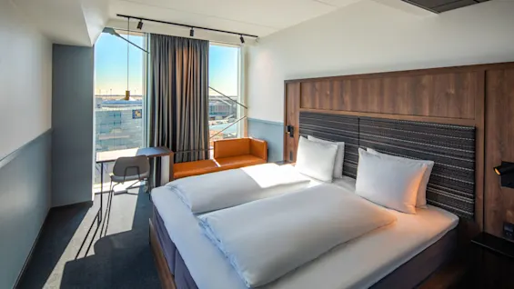 En pent, oppredd dobbeltseng står i et moderne hotellrom med stort vindu, sittegruppe, skrivebord, stol og utsikt over byen i sterkt dagslys.